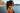 Woman with sunglasses and a scarf standing by a lake with mountains in the background